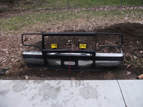 1990 Ford Bronco - A front bumper assembly with a black brush guard and two yellow fog lights is shown resting on the ground.