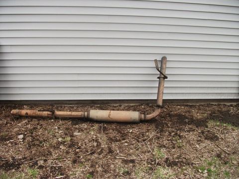 1990 Ford Bronco - An exhaust pipe and muffler assembly rests on the ground against a white-sided building.