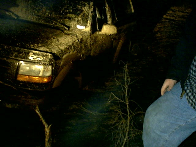 Muddy dark SUV at night, headlight lit, person in jeans nearby