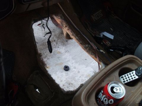 1990 Ford Bronco - The interior floor of a vehicle shows exposed metal and wiring, with a cut-out section and a soda can in the console.