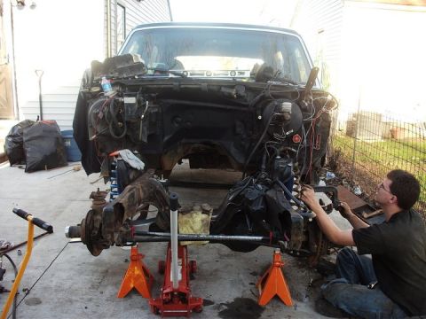 1990 Ford Bronco - A person works on the front suspension and engine bay of a disassembled Ford truck, which is elevated on jack stands.