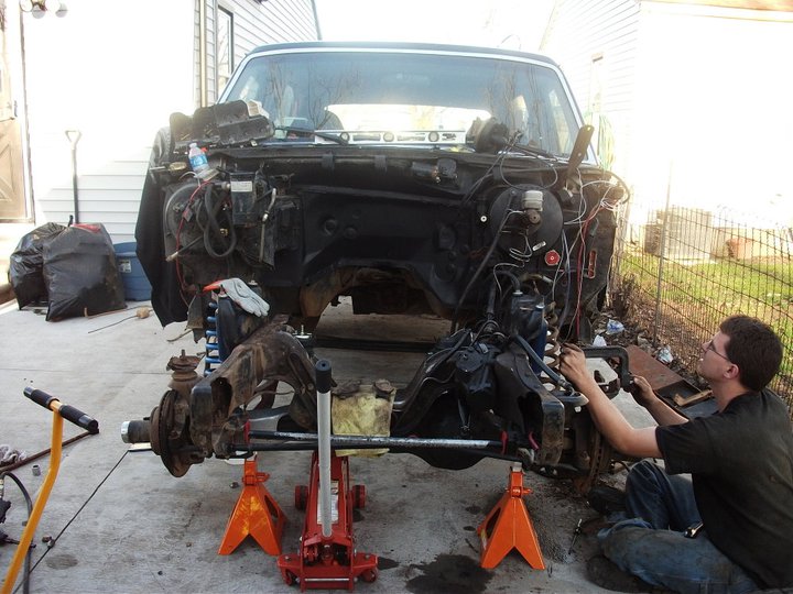 Ford truck body on jack stands with front end disassembled, a person works on the chassis