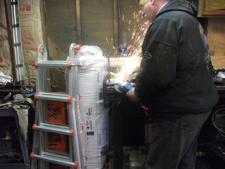 Man in garage using an angle grinder, sparks flying. Ladder and large bag visible