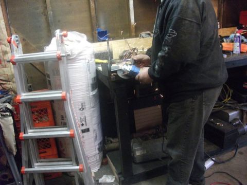 1990 Ford Bronco - A person is using an angle grinder in a workshop setting, with a ladder and a large bag visible.