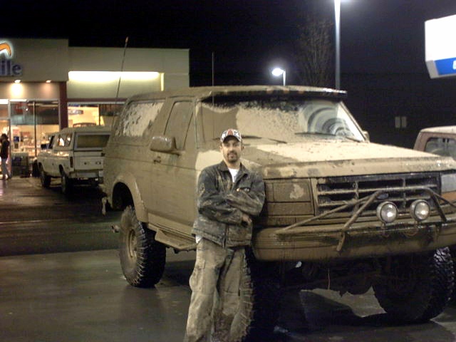Man in coveralls standing by a mud-covered lifted Ford Bronco SUV at night