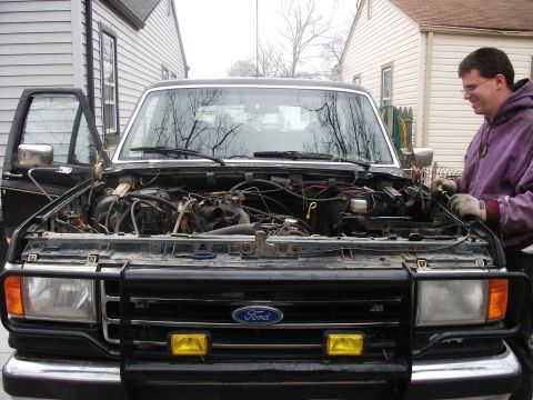 1990 Ford Bronco - A man works on the engine of a black Ford Bronco, which is parked in a driveway with houses in the background.