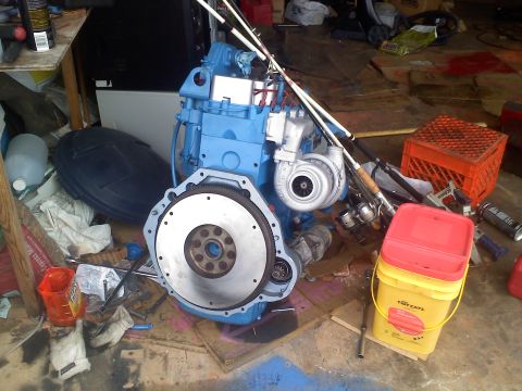 1990 Ford Bronco - A blue engine assembly with a turbocharger and flywheel is surrounded by fishing gear and various containers in a cluttered garage.