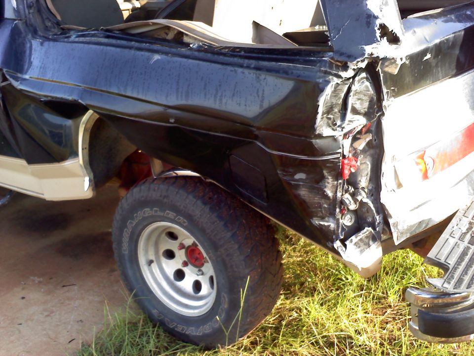 Rear corner of a black SUV with significant damage to the body and taillight area. The tire is visible