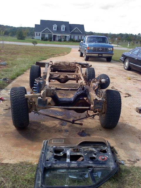 1995 Ford Bronco - A disassembled vehicle chassis sits on a driveway, with a blue pickup truck and a black sedan visible in the background.