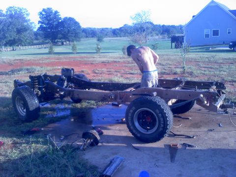 1995 Ford Bronco - A person works on the chassis of a vehicle outdoors, with a house and trees in the background.