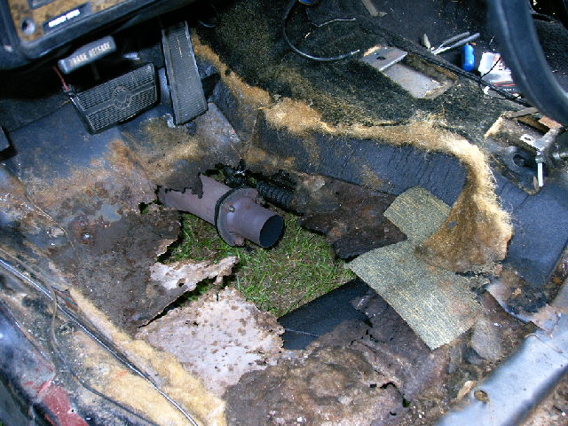 rusted car interior floor showing exposed grass and an exhaust pipe section