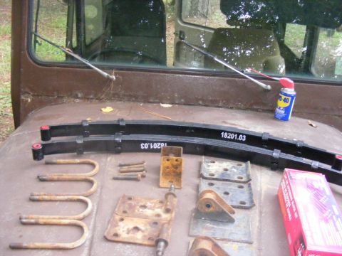 1978 Ford Bronco - Two black leaf springs with red bushings are laid out on a rusty brown surface, alongside U-bolts, bolts, and some metal brackets. A can of…