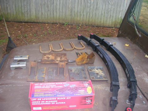 1978 Ford Bronco - A collection of vehicle suspension parts, including leaf springs, U-bolts, and lift blocks, are laid out on a rusty surface, possibly the h…