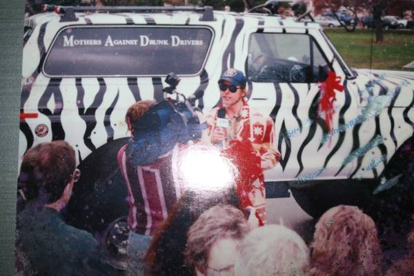 Man with microphone in front of a zebra-striped SUV with "Mothers Against Drunk Drivers" text.