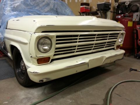 1967 Ford F-100 - The front end of a cream-colored pickup truck is shown, covered partially with blue plastic sheeting. The vehicle features a large horizont…
