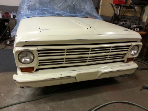 1967 Ford F-100 - The front end of a cream-colored 1967 Ford F-100 pickup truck is shown, covered in a blue tarp. The truck is in a workshop setting, with to…