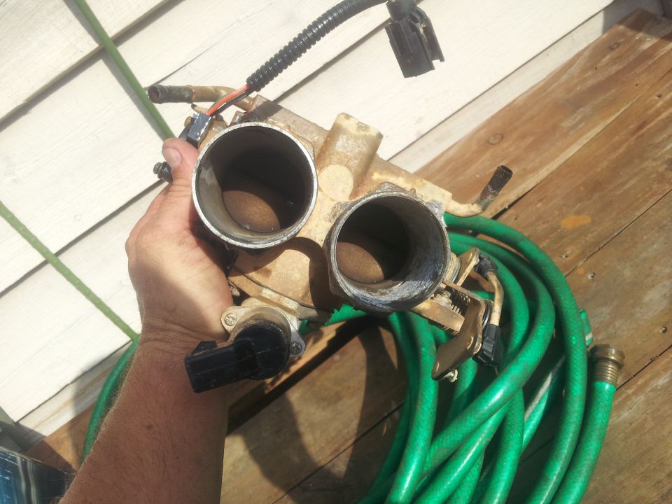 Hand holding a dirty dual throttle body assembly with a green garden hose coiled nearby