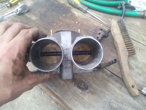 1988 Ford Bronco - A dirty dual throttle body is held in a hand, with various tools and a wire brush nearby on a workbench.