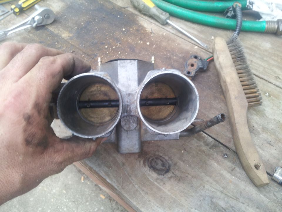 Hand holding a dual throttle body with a wire brush and tools on a wooden surface