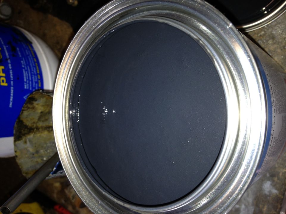 Open can of dark gray paint with a metal lid rim. A blue and white bucket is visible to the left