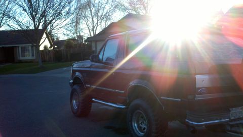 1992 Ford Bronco - A dark-colored Ford Bronco is parked on a residential street during sunset, with the sun's rays creating a lens flare.