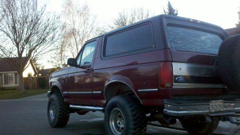 1992 Ford Bronco - A maroon 1992 Ford Bronco is parked on a residential street, showing its lifted suspension and spare tire on the back.