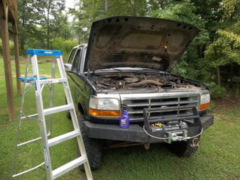 1995 Ford Bronco - A dark-colored Ford truck sits outside with its hood raised, revealing the engine bay. A ladder is positioned to the left of the vehicle.