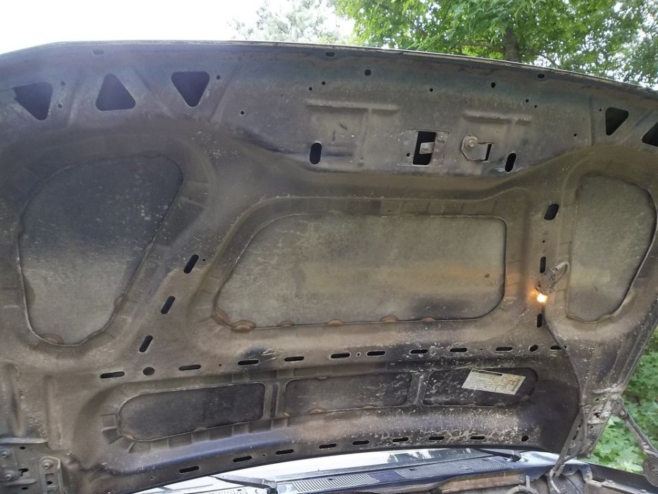 Underside of a dark blue car hood, showing insulation and latch mechanisms. A small sticker is visible
