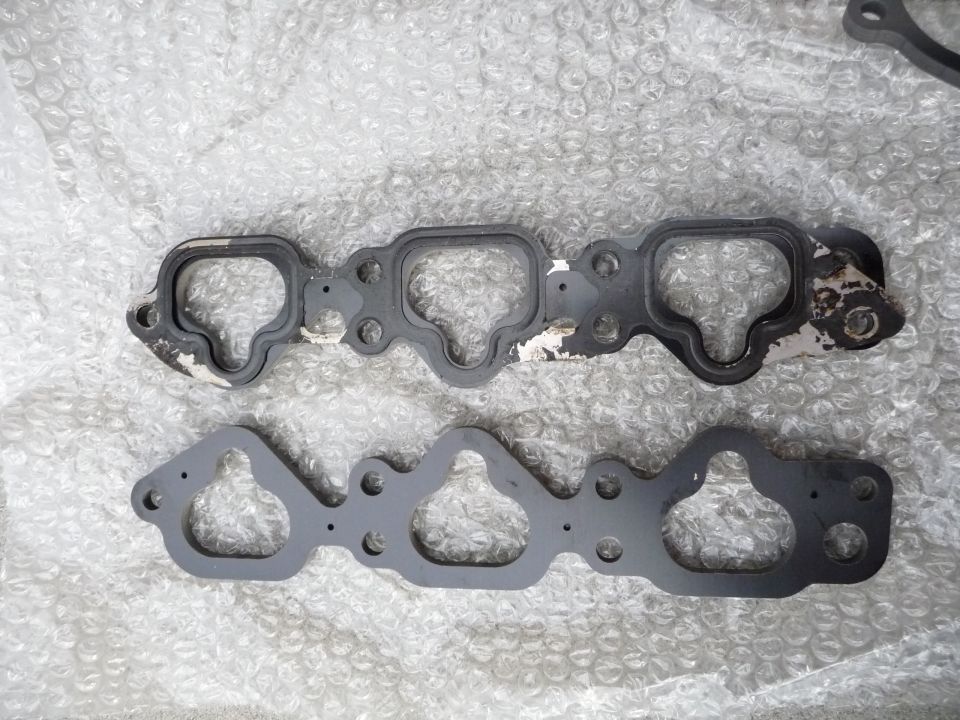 Two engine gaskets on bubble wrap. One gasket is black with white peeling paint and a black rubber seal. The other is dark gray