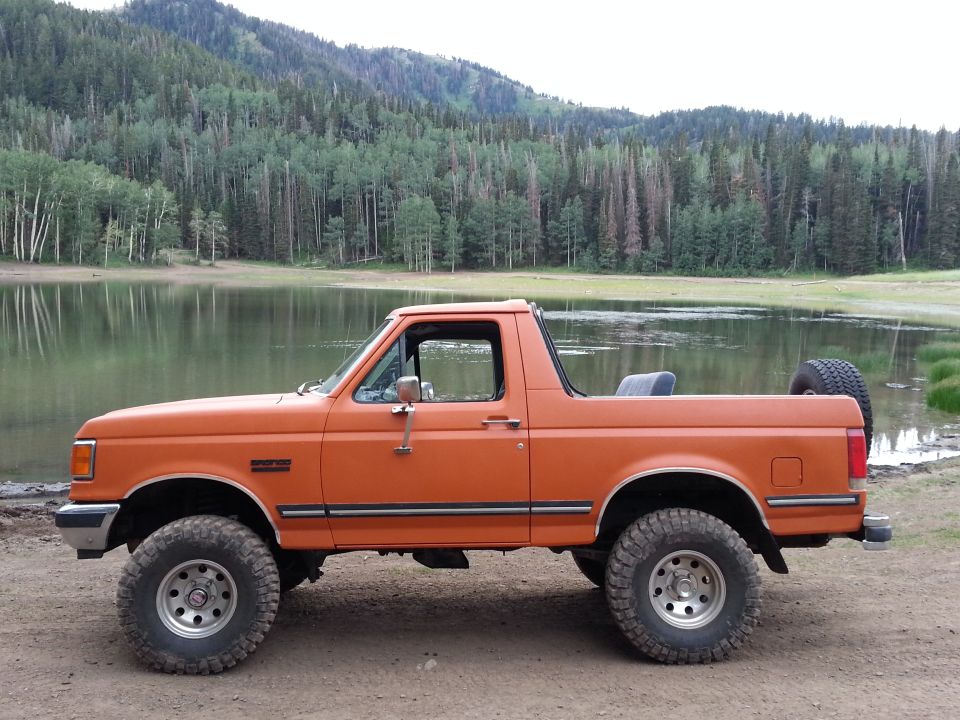 Side view of an orange Ford Bronco parked by a lake with a mountain backdrop.