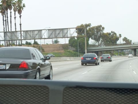 2003 Ford Crown Victoria - Black sedans drive a freeway beneath a steel overhead sign truss, with palm trees lining the left shoulder.