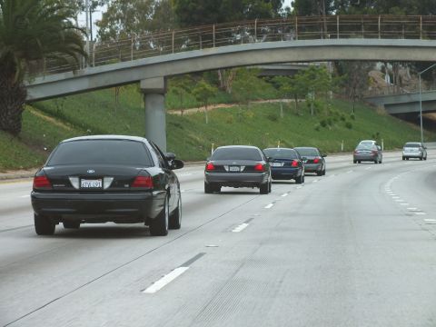 2003 Ford Crown Victoria - procession of black Ford sedans follows one another along a curving freeway beneath a concrete overpass.