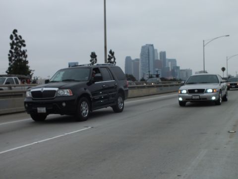 2003 Ford Crown Victoria - Photograph of a black Lincoln Navigator and a silver sedan driving on a freeway with a city skyline behind.