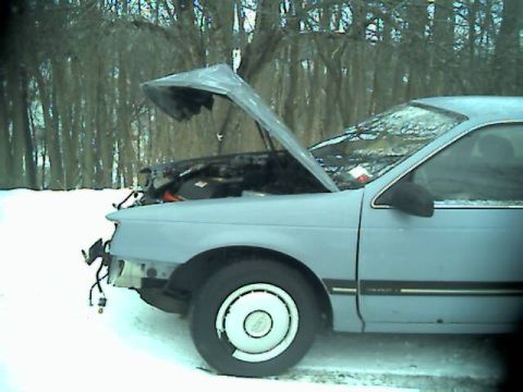 1986 Ford Taurus - A light blue 1986 Ford Taurus sits in the snow with its hood open, showing signs of damage to the front bumper.