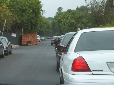 2003 Ford Crown Victoria - Photo of a line of cars driving down a tree-lined street past a rusty rental dumpster.