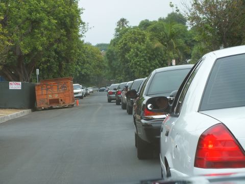 2003 Ford Crown Victoria - Photo of a convoy of cars on a shaded residential street beside a rusty rental dumpster.