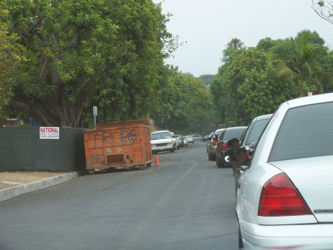 2003 Ford Crown Victoria - Photo of cars lined along a newly paved street beside a graffitied rental dumpster.