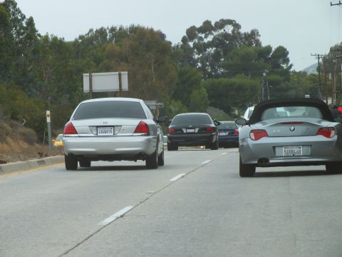 2003 Ford Crown Victoria - Road photo of a silver sedan and a roadster on a two-lane road with trees behind, not a technical document.