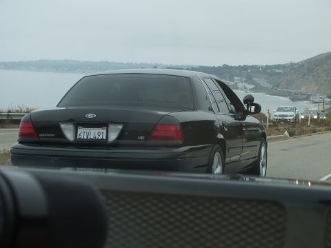 2003 Ford Crown Victoria - Road photo of a black Ford Crown Victoria rear with a California plate by a coastline, not a technical document.
