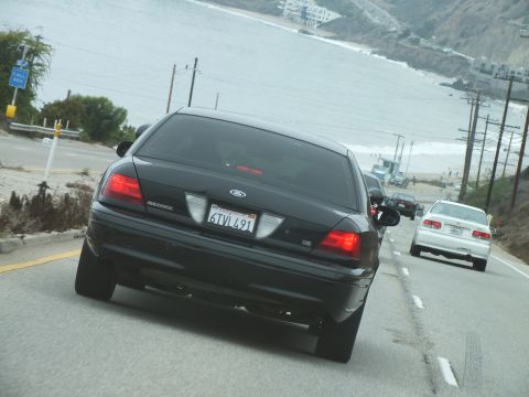 2003 Ford Crown Victoria - Driving photo of a black Ford Crown Victoria rear on a coastal highway near a beach, not a technical document.
