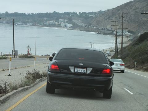 2003 Ford Crown Victoria - Driving photo of a black Ford Crown Victoria rear on a coastal highway above the ocean, not a technical document.
