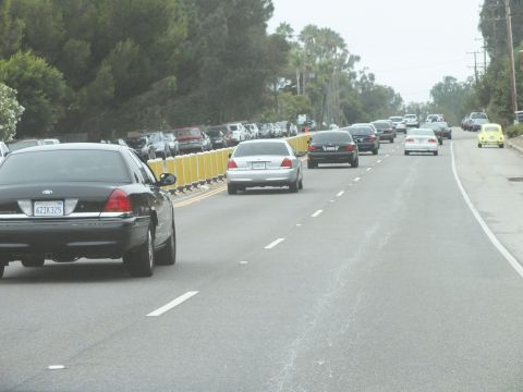 2003 Ford Crown Victoria - Black and silver sedans travel a divided road lined with yellow barrier posts and dense roadside trees.