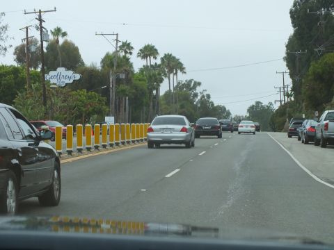 2003 Ford Crown Victoria - silver sedan travels past parked cars and a Geoffrey's sign on a coastal road lined with yellow barrier posts.