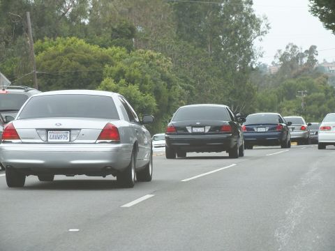 2003 Ford Crown Victoria - silver Ford Crown Victoria and a black sedan are seen from behind on a road bordered by dense green trees.