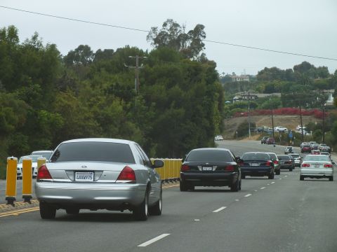 2003 Ford Crown Victoria - silver Ford Crown Victoria leads dark sedans on a road with yellow barrier posts and distant green hills.