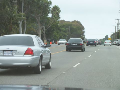 2003 Ford Crown Victoria - Photograph of a silver Crown Victoria and black sedans on a tree-lined coastal highway, not a technical document.