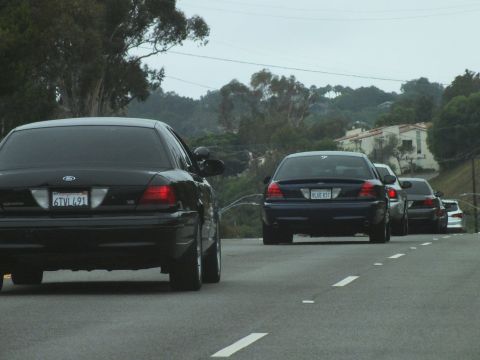 2003 Ford Crown Victoria - Photograph of a black Crown Victoria and dark sedan on a coastal highway, not a technical document.