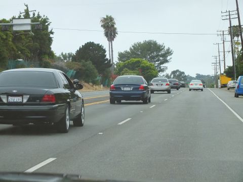 2003 Ford Crown Victoria - Photograph of a black Crown Victoria and blue sedan on a multi-lane coastal highway, not a technical document.