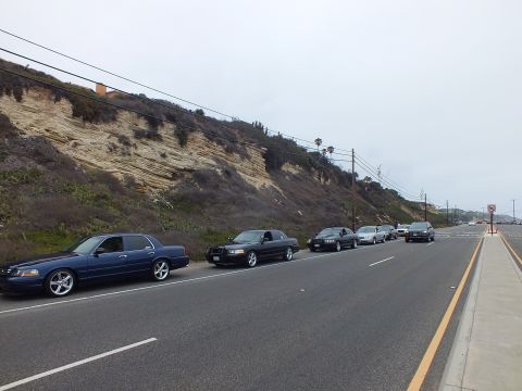2003 Ford Crown Victoria - Photograph of a row of dark sedans parked along a coastal highway shoulder, not a technical document.