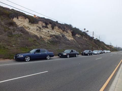 2003 Ford Crown Victoria - Photograph of several sedans parked nose-to-tail along a coastal highway, not a technical document.